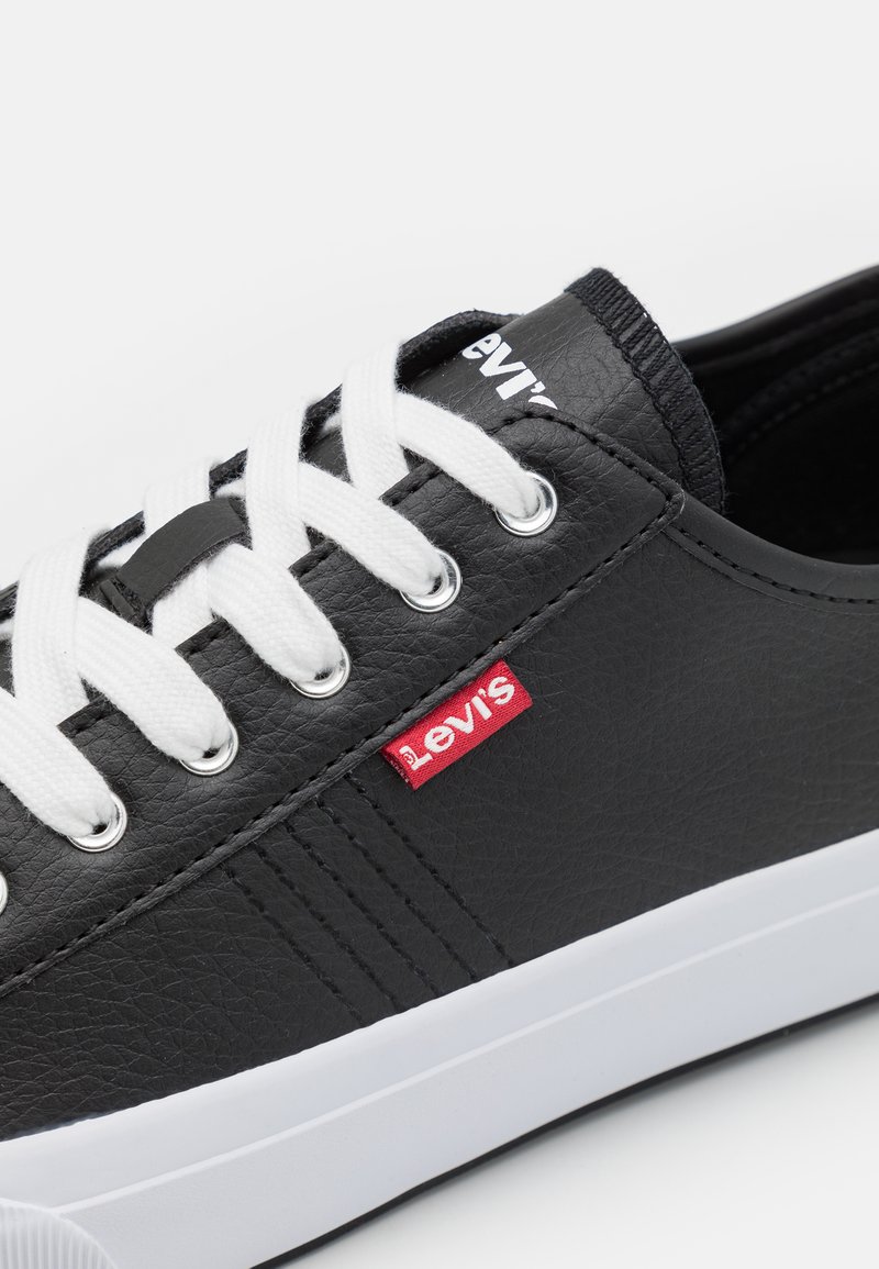 Zapatilla de piel negra con cordones blancos y suela de goma. Presenta una etiqueta roja con el logo de Levi's y ojales plateados. Superficie texturizada con detalles de costura.