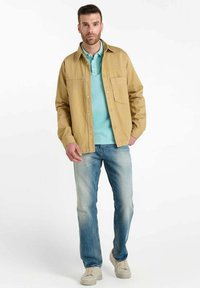 Veste en coton brun clair sur un polo turquoise, associée à un jean bleu délavé et des baskets beiges. Tissu texturé avec un détail de poche à l'avant.