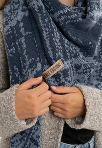 Écharpe tricotée en bleu marine et bleu clair avec un motif texturé. L'étiquette indique "Knit Factory". Les mains du modèle tiennent l'écharpe, portant un pull gris.