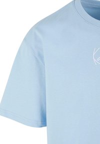 Light blue cotton t-shirt with short sleeves and a crew neckline, featuring a white embroidered logo on the chest. Smooth texture, casual design.
