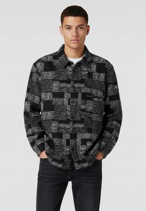 Camicia a scacchi nera e grigia con design a bottoni, tessuto texturizzato e taschino sul petto, abbinata a jeans scuri.