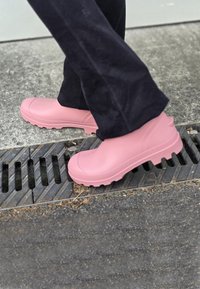 Sabots en caoutchouc rose avec une finition lisse et une semelle texturée, dotés d'un bout arrondi et de bords surélevés, portés avec un pantalon noir sur un pavé.