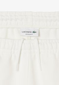 Pantalon de survêtement blanc avec une taille élastique à cordon de serrage, une texture douce, et une étiquette affichant "LACOSTE" et "COUPE RÉGULIÈRE."