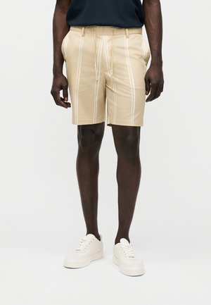 Muškarac u bež kratkim hlačama s bijelim vertikalnim prugama, crnoj košulji i bijelim tenisicama, stoji na jednobojnoj bijeloj pozadini.