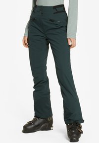 Pantalones de esquí verdes hechos de material flexible, con una cintura alta, bolsillos con cremallera y puños reforzados. Llevados con botas de esquí.