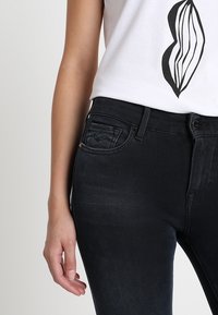 Mustat denim-farkut, joissa on kapea istuvuus, pieni etutasku ja hillityt ompelut yksityiskohdat. Yhdistetty valkoiseen paitaan, jossa on graafinen kuvio.