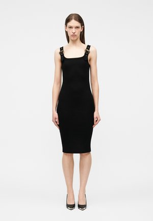 Mujer de pie vestida con un vestido negro hasta la rodilla con tirantes anchos y detalles de hebilla dorada, combinado con zapatos de tacón altos negros y puntiagudos.