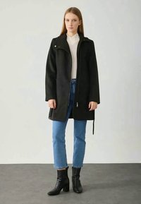 Cappotto oversized nero con chiusura a zip, tasche laterali e un dettaglio con bottone a pressione sul colletto, abbinato a jeans azzurri chiari e stivali neri alla caviglia.