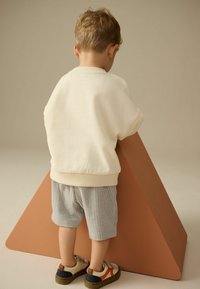 Enfant portant un sweat-shirt crème et un short à rayures bleues, s'appuyant contre une structure triangulaire marron, chaussé de baskets multicolores.