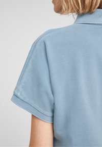 Vaaleansininen polo-paita, lyhyet hihat, teksturoitu kangas ja tasakudottu kaulus. Pistoiluseikat näkyvät olka-sauman lähellä.