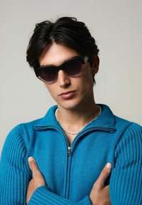 Des lunettes de soleil noires en forme d'œil de chat avec des verres dégradés, portées par une personne dans un pull zippé côtelé teal. Le pull présente un col haut et des poignets texturés.