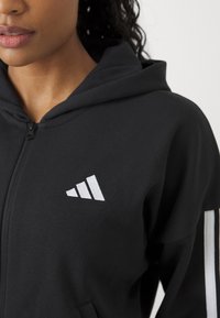Svart zip-up hoodie i mjukt material, med en vit logotyp med tre ränder på bröstet och vita sidostripes på ärmarna.