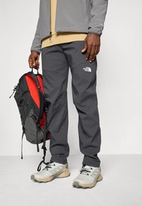 Grijze buitenbroek met een wit logo, gecombineerd met een grijze jas, beige shirt en grijze sneakers, terwijl hij een zwarte en rode rugzak vasthoudt.