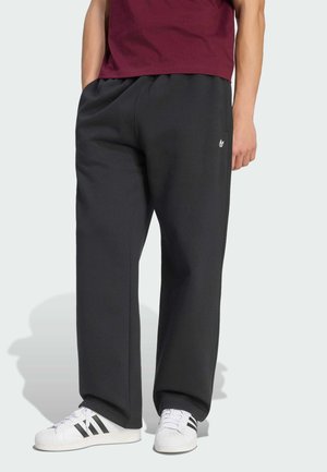 Person iført løse sorte Adidas joggingbukser med hvidt logo, hvide sneakers med sorte striber og en vinrød skjorte, der står med hænderne i lommerne.