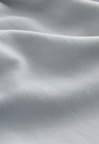 Lichtgrijze stof met een gladde, matte textuur en subtiele glans, zacht vallend in zachte plooien, die het zachte, synthetische materiaal benadrukt.