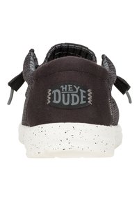 Sneaker nera con tomaia testurizzata, etichetta sul tallone con la scritta "HEY DUDE" e suola in gomma bianca maculata. Dettagli includono lacci piatti e occhielli.