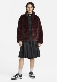 Mörkröd fuskpälsjacka med dragkedja, svart vågmönster; matchad med en svart plisserad kjol och sneakers i vitt och svart, med rosa strumpor.