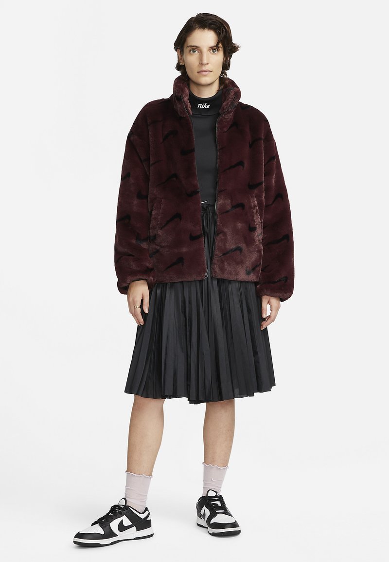 Veste en fausse fourrure bordeaux avec fermeture éclair, motif vague noir ; associée à une jupe plissée noire et des baskets blanc-noir, avec des chaussettes roses.
