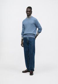 Gaiši zils teksturēts džemperis pār pelēku augstu apkaklīti, saskaņots ar platajiem ziliem džinsiem un tumši brūniem loaferiem. Vienkārša un ikdienišķa apģērba kombinācija.