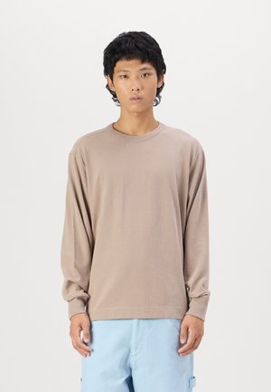 Beige pamut hosszúujjú póló, kerek nyakkivágással, laza szabással, sima textúrával. Világoskék nadrággal párosítva, laza kialakítással.