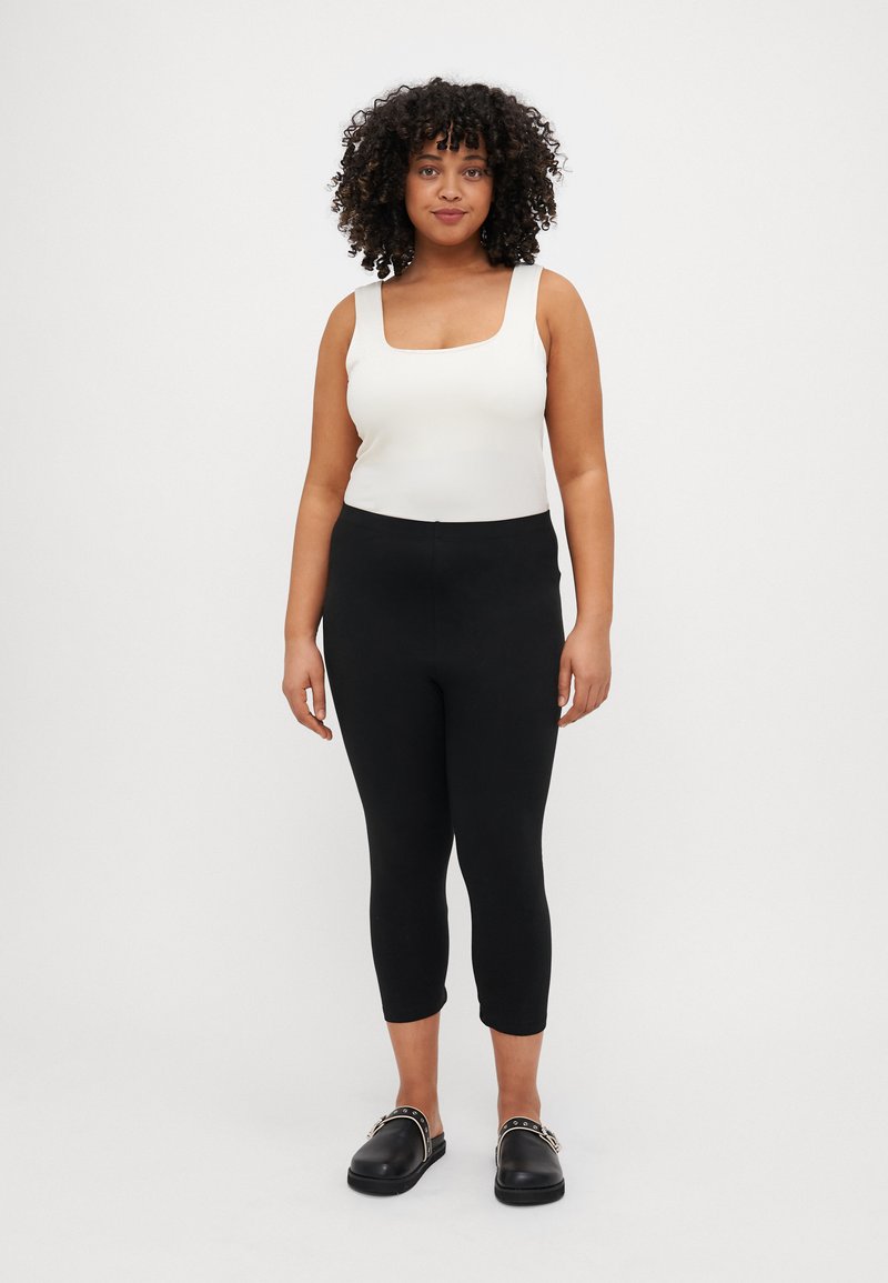 Black leggings with a stretchy material, paired with a white square-neck tank top, and black slip-on shoes with silver accents.