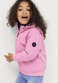 Rosa Softshell-Jacke mit Kapuze, Reißverschluss und elastischen Bündchen. Mit einem schwarzen runden Logo-Patch am linken Arm.