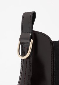 Botín de cuero negro con textura suave, que presenta un aro de metal dorado en el tobillo y costuras en marrón contrastante a lo largo de las costuras.