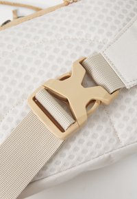 Beige stoffen gesp met een ingewikkeld stippenpatroon, voorzien van een tan kleurige clip en een geweven band, met textuurdetails en robuuste hardwaren.