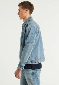 Chaqueta de denim azul clara con un diseño clásico, que presenta un bolsillo en el pecho, cierre de botones y detalle de costuras a lo largo de las costuras.