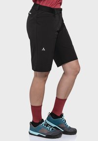 Schwarze Outdoor-Shorts mit lockerer Passform, ausgestattet mit Taschen und einem Logo auf dem Oberschenkel, kombiniert mit blauen und grauen Wanderschuhen und roten Socken.