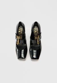 Zapatos Mary Jane de charol negro con punta cuadrada, que presentan una hebilla decorativa dorada y la marca Versace en la plantilla.