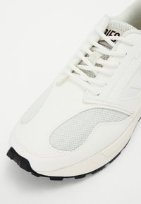 Baskets blanches de sport avec panneaux en mesh et lacets, dotées d'une semelle noire texturée et d'une languette matelassée avec un marquage subtil.