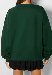 Skoggrön oversized sweatshirt med sänkt axel, ribbad hals och ärmslut, i kombination med en plisserad svart minikjol. Mjuk tygtextur.