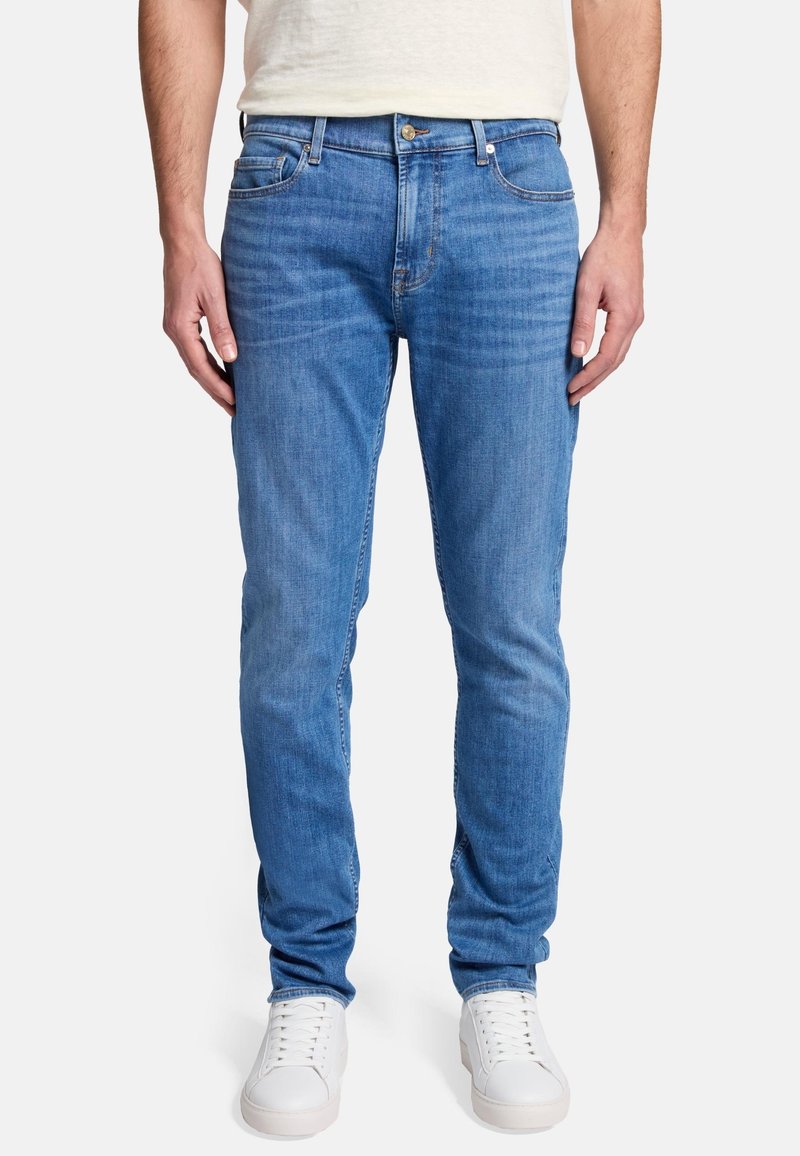 Jean en denim bleu clair au design ajusté, cinq poches, couture discrète et fermeture à boutons à la taille. Portés avec des baskets blanches.