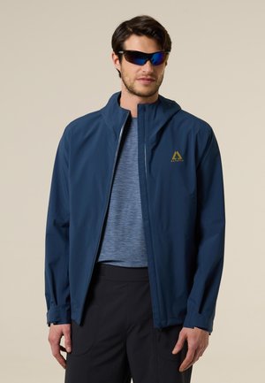 Homme portant une veste Altavia bleue, une chemise grise, un pantalon noir et des lunettes de soleil enveloppantes réfléchissantes sur un fond uni.