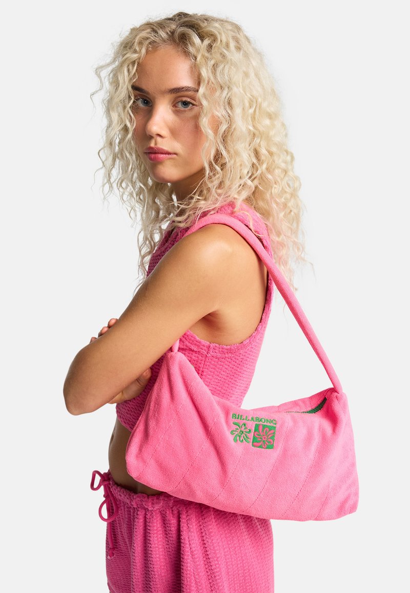 Jonge vrouw met krullend blond haar draagt een roze gestructureerde mouwloze top en korte broek, met een roze Billabong schoudertas.