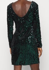 Černé a tmavě zelené šaty s flitry, s hlubokým V-výstřihem na zádech, dlouhými rukávy a přiléhavým střihem, které předvádějí lesklou texturu a vertikální vzor.