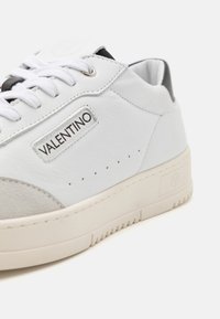 Valkoinen nahkainen Valentino-tenniskenkä valkoisilla nauhoilla, beige nupukkinokalla, mustalla kantapääyksityiskohdalla sekä brändin logolla sivulla ja pohjassa.