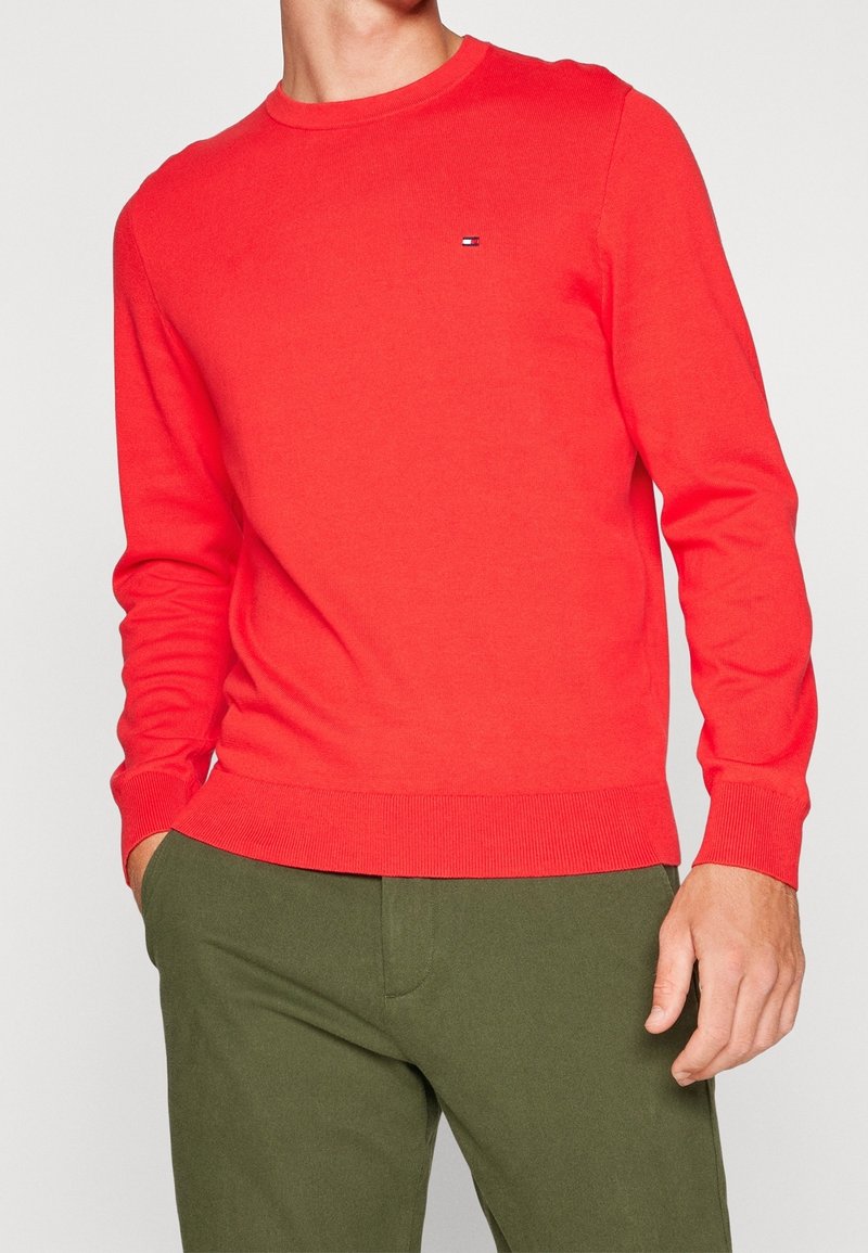 Pull rouge en maille avec manches longues et poignets côtelés, arborant un petit logo sur la poitrine. Assorti à un pantalon vert olive.