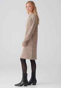 Abito lavorato a maglia beige fino al ginocchio con finitura testurizzata, maniche lunghe e scollatura rotonda, abbinato a stivali neri alla caviglia e collant.