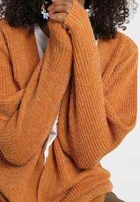 Cárdigan naranja de punto con mangas largas, textura acanalada y corte holgado, superpuesto a una camisa blanca. Mangas completas.