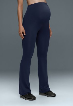 Personne enceinte portant des leggings de maternité bleu marine et des chaussures de sport noires, debout devant un fond gris.