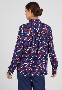 Camicia a maniche lunghe in tessuto fantasia con motivi geometrici viola, blu e bianchi. Con colletto e dettaglio a pieghe sul retro.