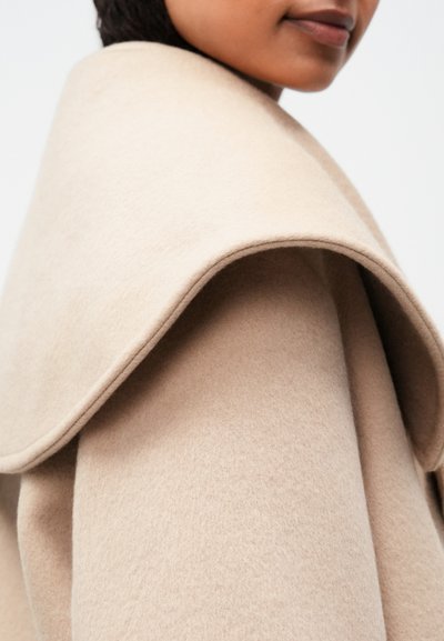 Beige gyapjúkabátot viselő személy, széles, hajtott gallérral, vállatól felfelé, egyszínű háttér előtt.