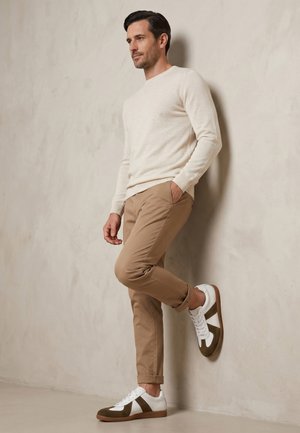 Muž opírající se o zeď, oblečený v krémovém svetru, béžových kalhotách a bílých a hnědých teniskách, s jednou rukou v kapse.