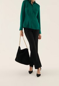 Blusa di seta verde con maniche lunghe e bottoni, abbinata a pantaloni neri a campana e una grande borsa a tracolla in suede nero con catena dorata.