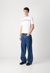 Hvid t-shirt med korte ærmer og sort tekst, parret med løstsiddende mørkeblå denimjeans og sorte sko. Simpelt design med en afslappet stil.