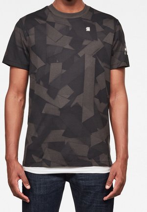 Mand iført en sort og mørkegrå t-shirt med geometrisk mønster og korte ærmer med en lille hvid logo på brystet og mørke jeans.