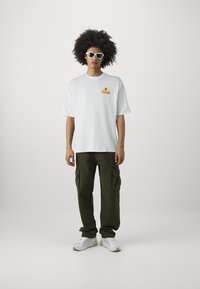 Camiseta blanca de manga corta con un gráfico de "FRESH" en naranja y amarillo. Pantalones cargo verde oliva con bolsillos laterales y zapatillas blancas.