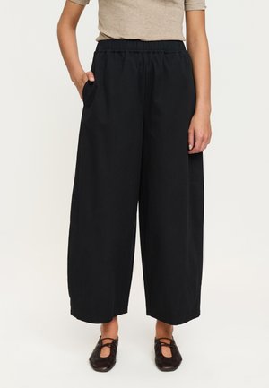 Persoon die losse zwarte wijde broek met elastische tailleband draagt en één hand in de zak, gecombineerd met bruine platte schoenen.